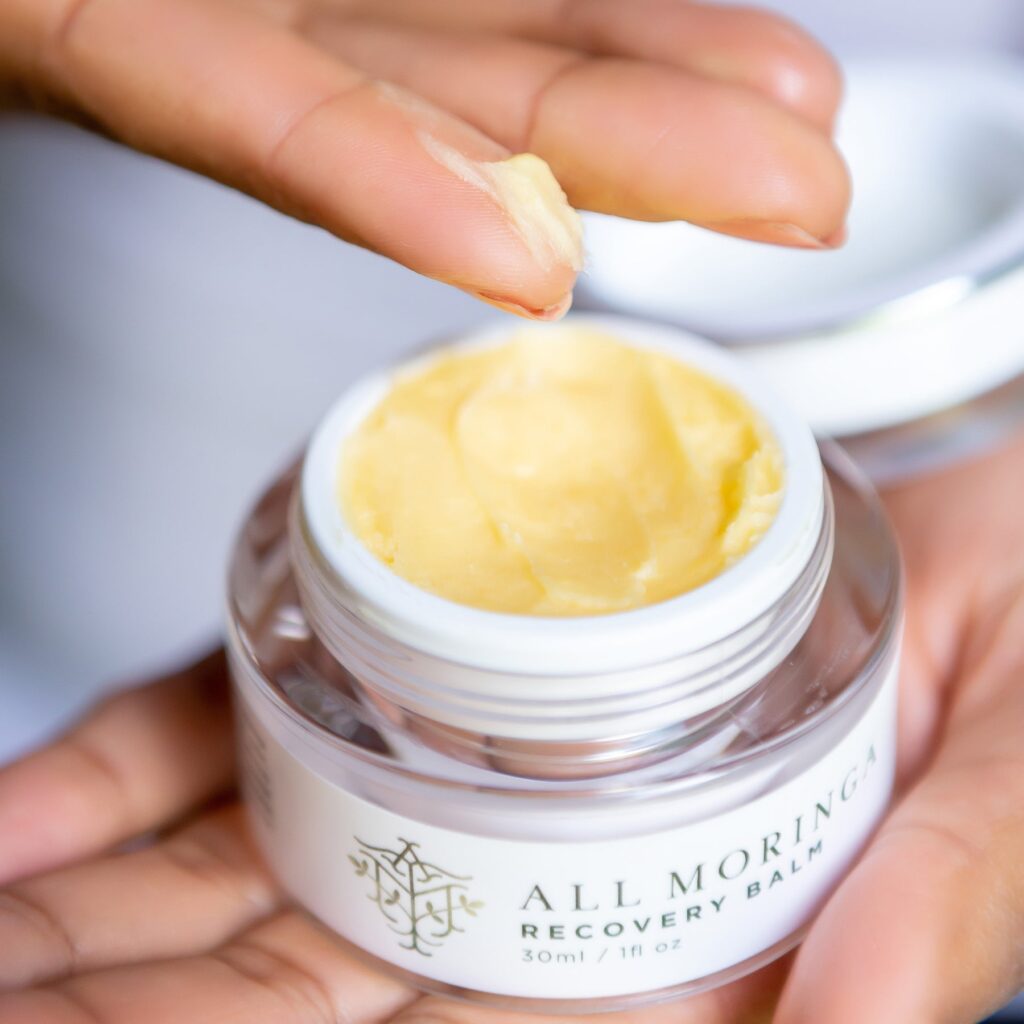 Moringa recovery balm held between fingers, showcasing its creamy texture. The jar is labeled for skincare benefits and natural healing.