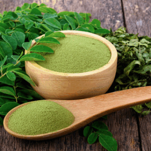 Green moringa powder in a wooden bowl and spoon, surrounded by fresh moringa leaves, highlighting its health benefits and culinary uses.