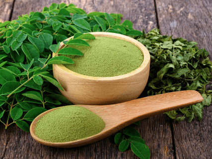 Green moringa powder in a wooden bowl and spoon, surrounded by fresh moringa leaves, highlighting its health benefits and culinary uses.