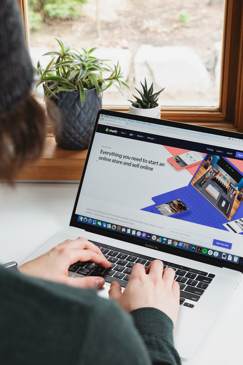 Person using a laptop on a table, viewing Shopify's website, promoting online store setup. Potted plants visible in a sunny window backdrop.